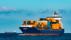A large blue cargo ship loaded with colorful shipping containers sailing across the open sea with another vessel in the background. Transportvibe
