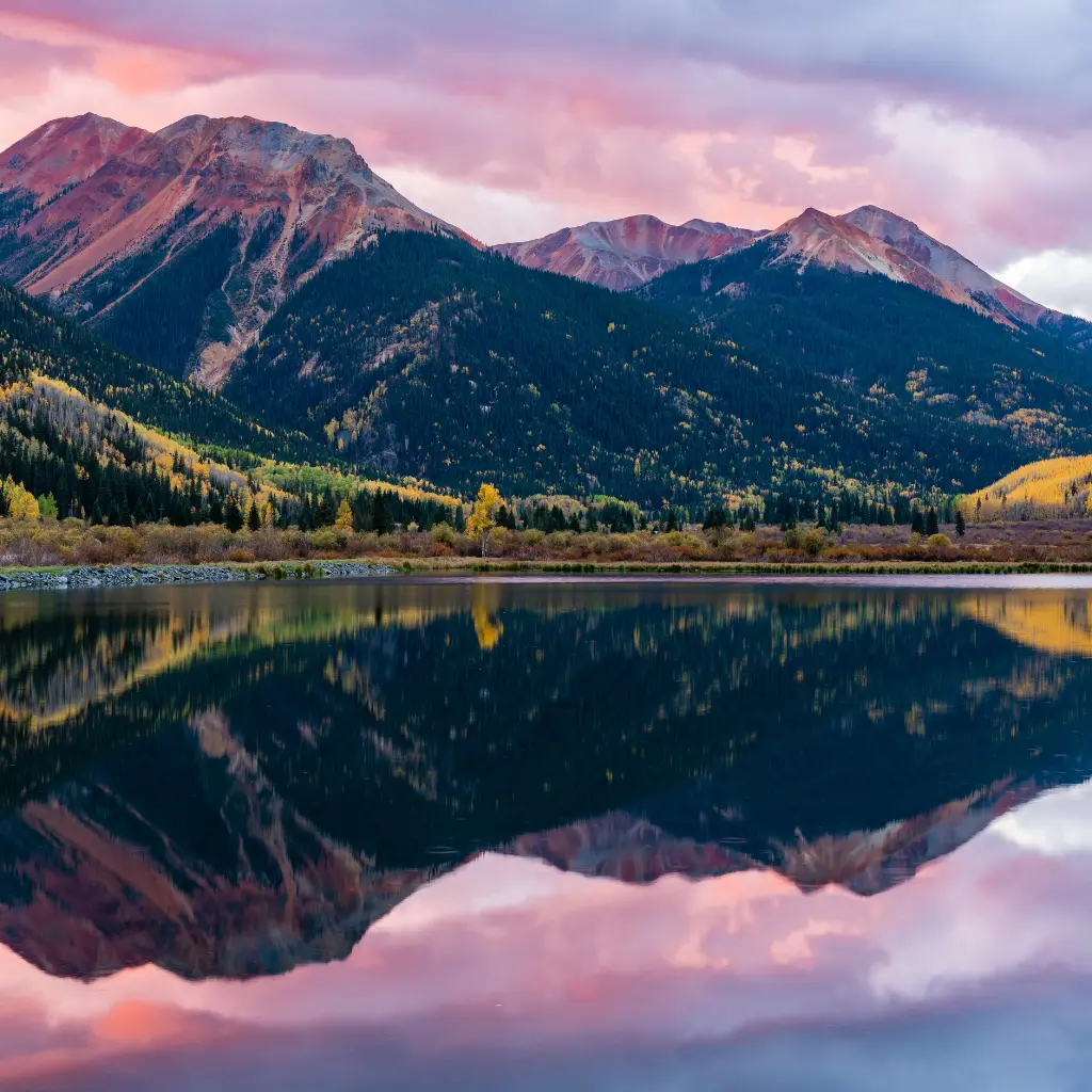 A breathtaking view of Colorado’s mountains at sunset, with vibrant autumn colors and a still lake reflecting the peaks and sky. Transportvibe