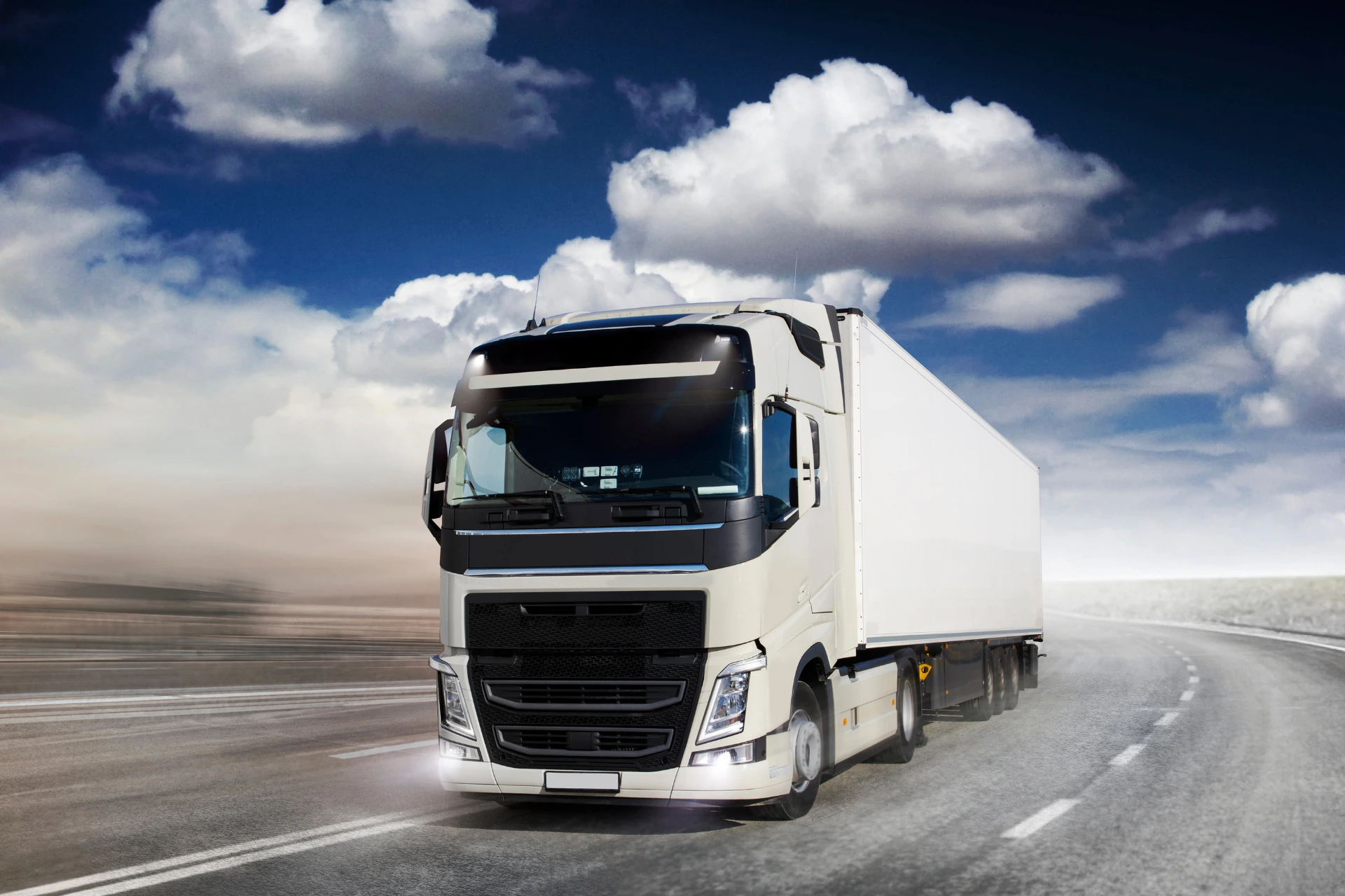A large white semi-truck is driving on an open highway under a dramatic cloudy sky, representing long-distance cross country car shipping.