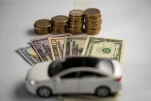 A small white car model on top of U.S. dollar bills, with stacks of coins in the background, representing the cost of a car shipping quote.